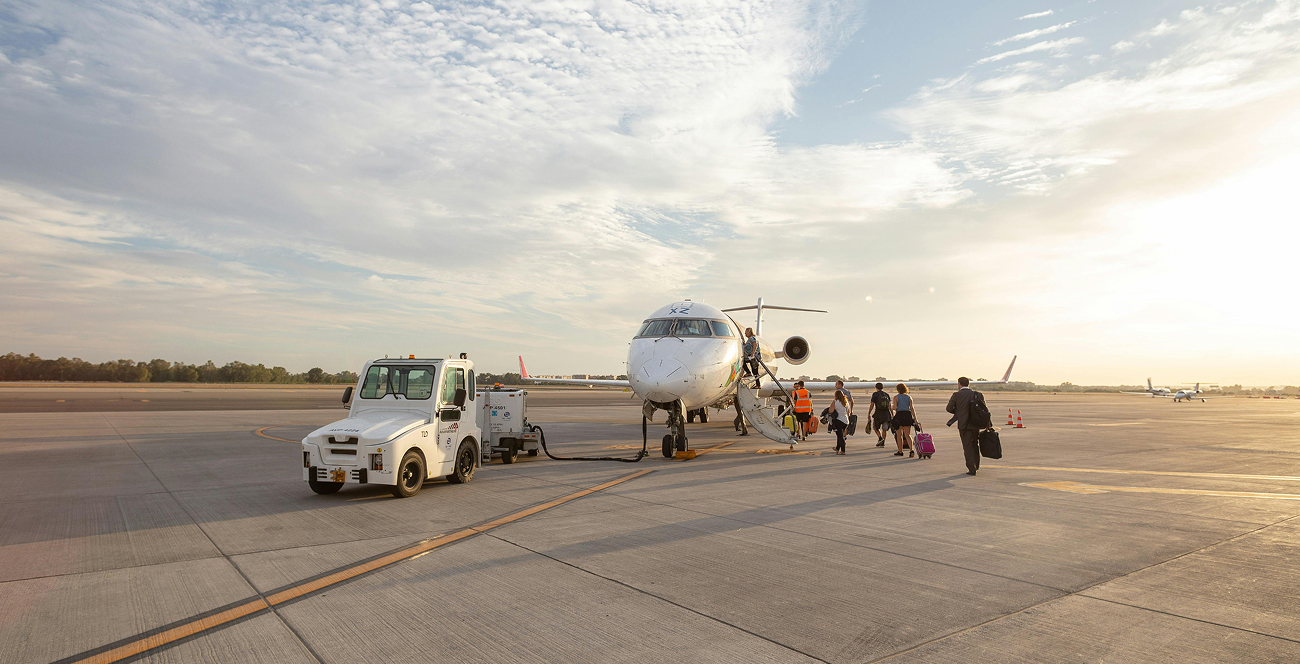 Resenärer går ombord på ett mindre passagerarflygplan på en flygplatsplatta i solnedgångsstämning, med ett markfordon parkerat intill.